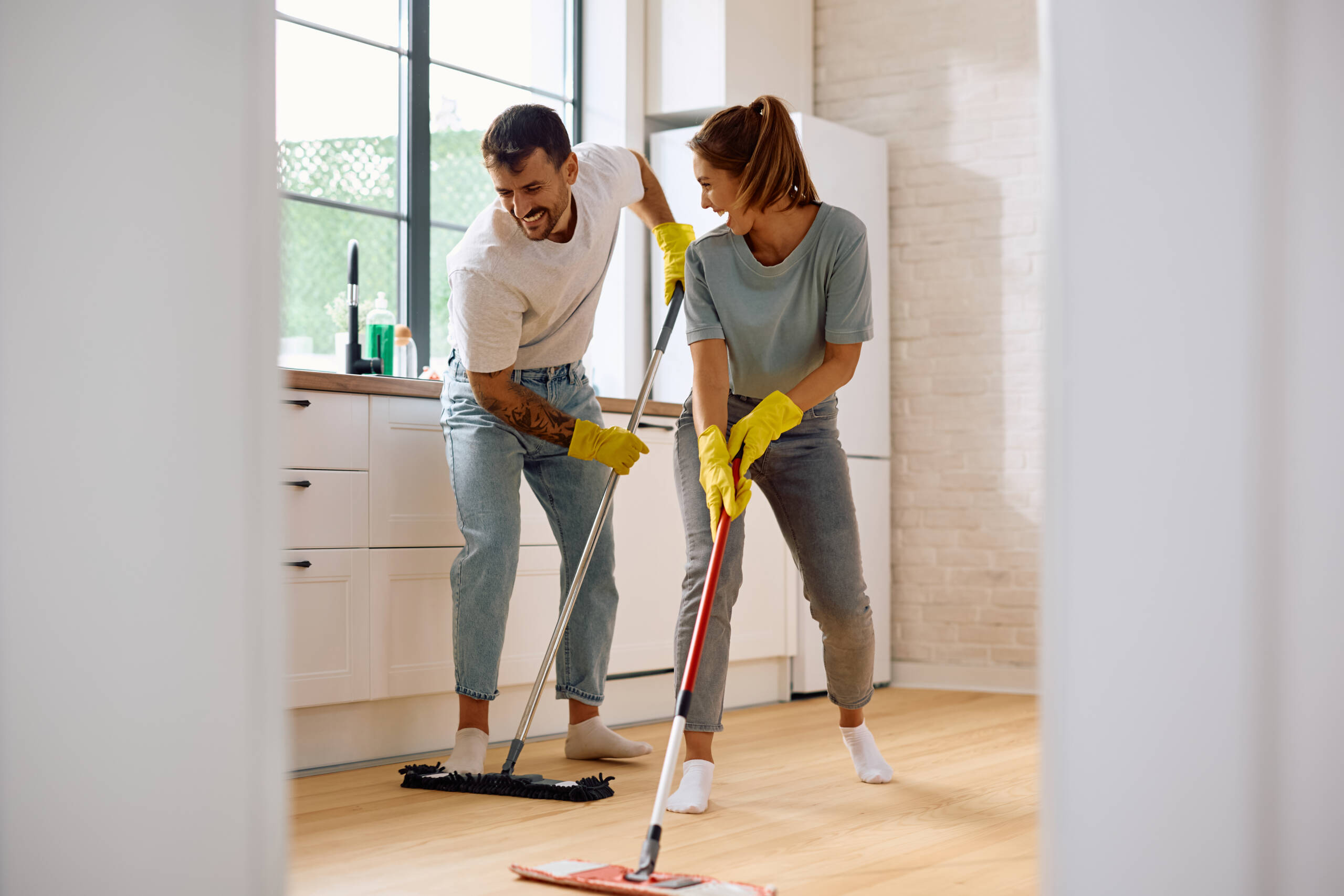 Young happy couple having fun while cleaning the floor int he kitchen with a mop. Copy space.